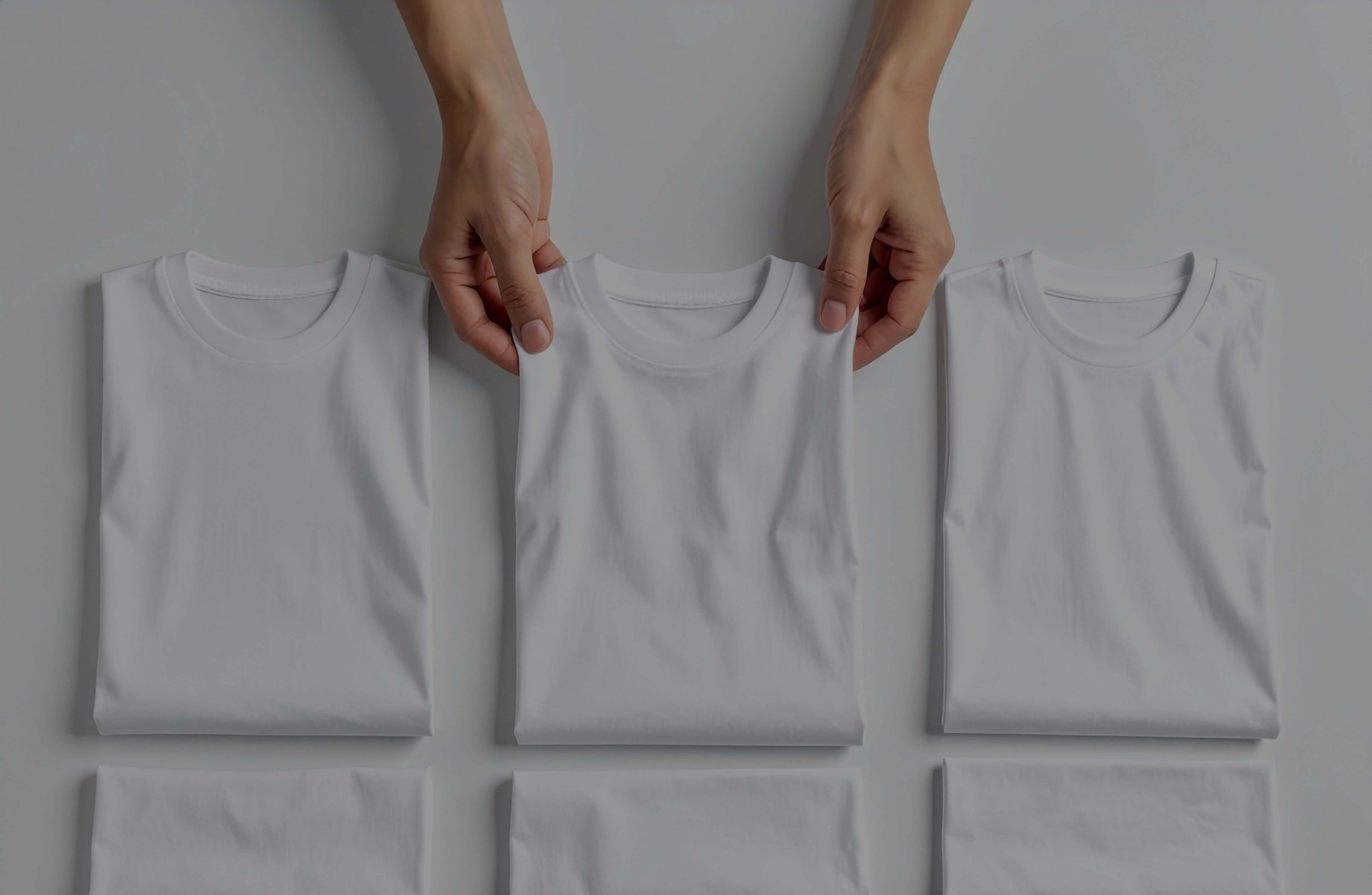 Drei weisse T-Shirts die in einer Reihe gelegt sind und eins wird von zwei Händen gehalten