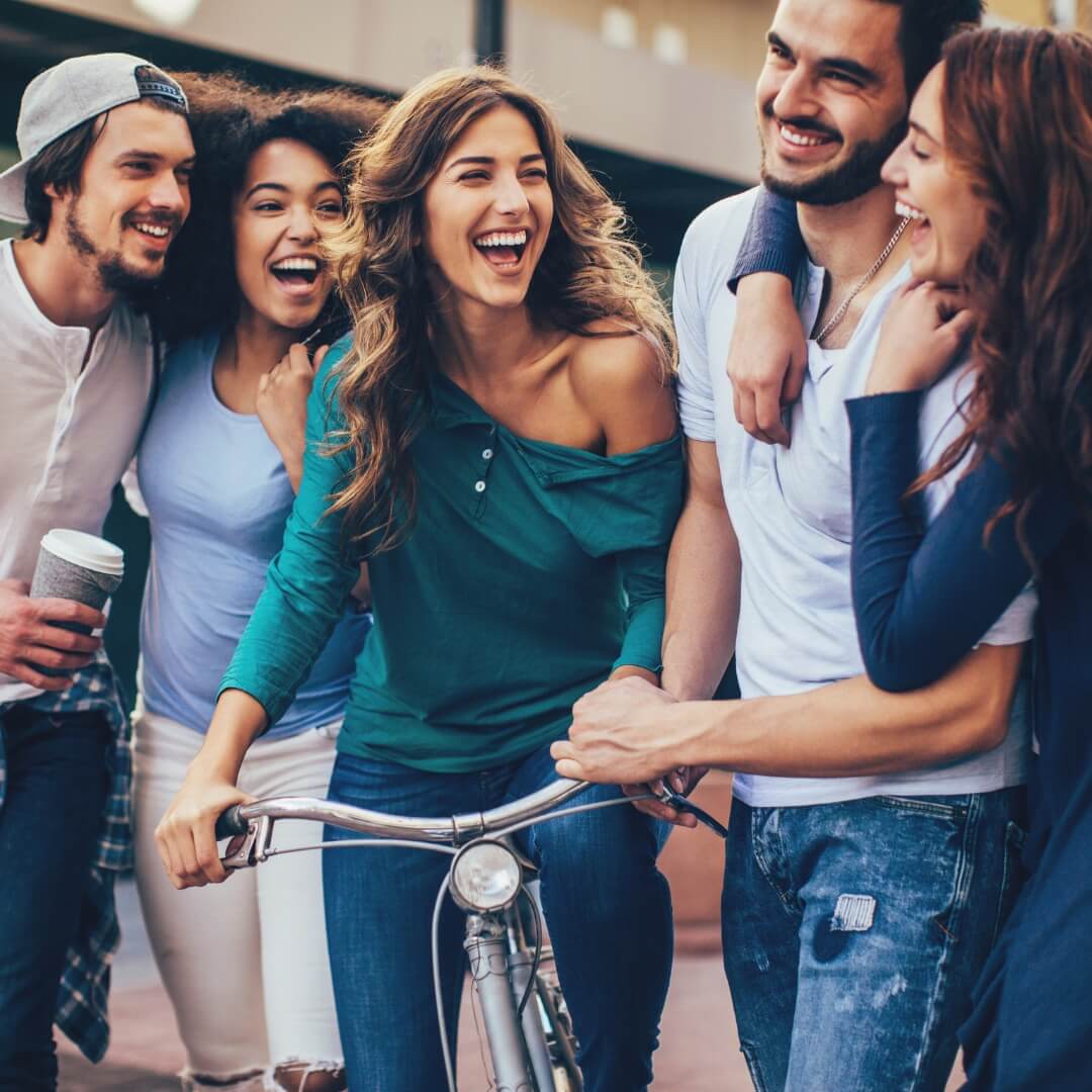 Eine Gruppe lachender junger Freunde in Freizeitkleidung steht zusammen im Freien; eine Frau sitzt auf einem silbernen Fahrrad, während ein Mann daneben einen Kaffeebecher hält.
